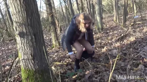 Blonde in dark jacket exposes legs while crouching in forest