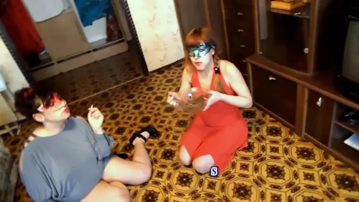 Two brunette women sit on floor, one eating while they talk and smoke